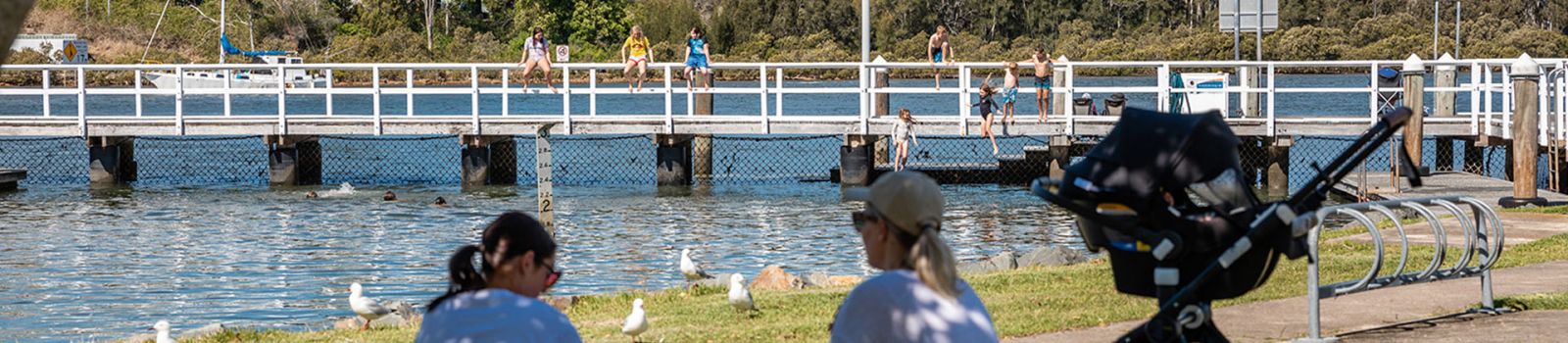 Image of parents sitting on the grass with a pram and overlooking a river and a walkway  banner image