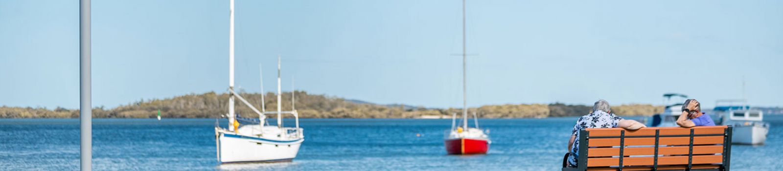 Image of 2 people sitting on a bench over looking a bay with yachts sailing past banner image