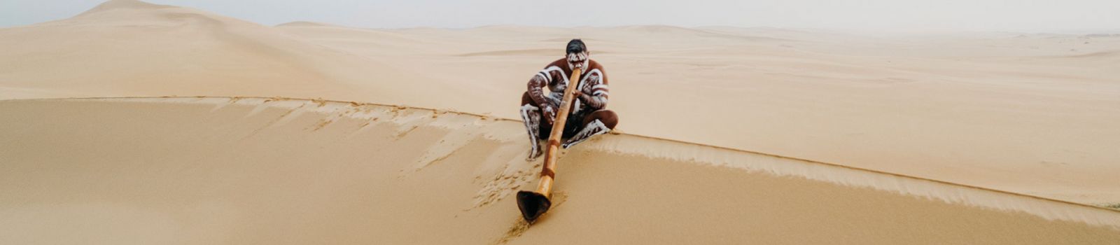 Image of an Aboriginal man playing a didgeridoo in the sand dunes banner image