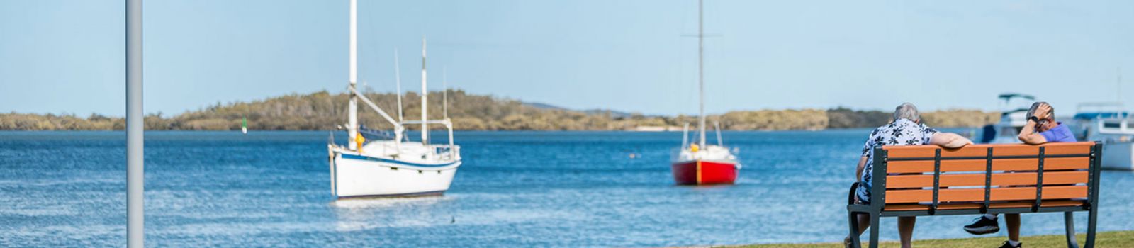 Image of two men sitting on a park bench overlooking a beach with yachts banner image